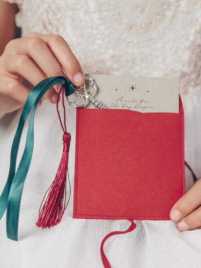 A child holds the silver key by its green ribbon beside a red suede pouch. The note card peeks out of the pouch.