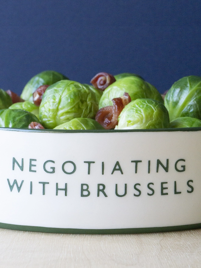 Ceramic porcelain dish with Negotiating with Brussels written on the side. A white Christmas serving dish with green rim containing Brussels sprouts and bacon.