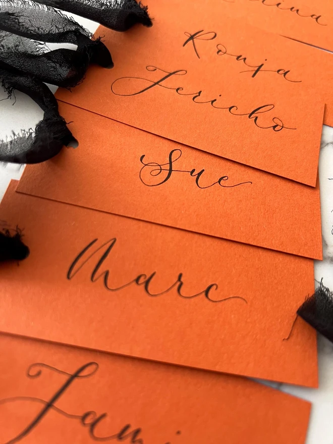 Personalised Calligraphy Rust Orange Place Card, orange place cards with black chiffon ribbons grouped together on a white surface. 