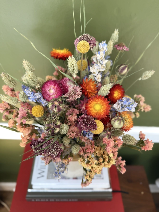 a dried flower bouquet of spring/summer colours in a victorian jam pot sitting on top of a stack of books and magazines against a green wall