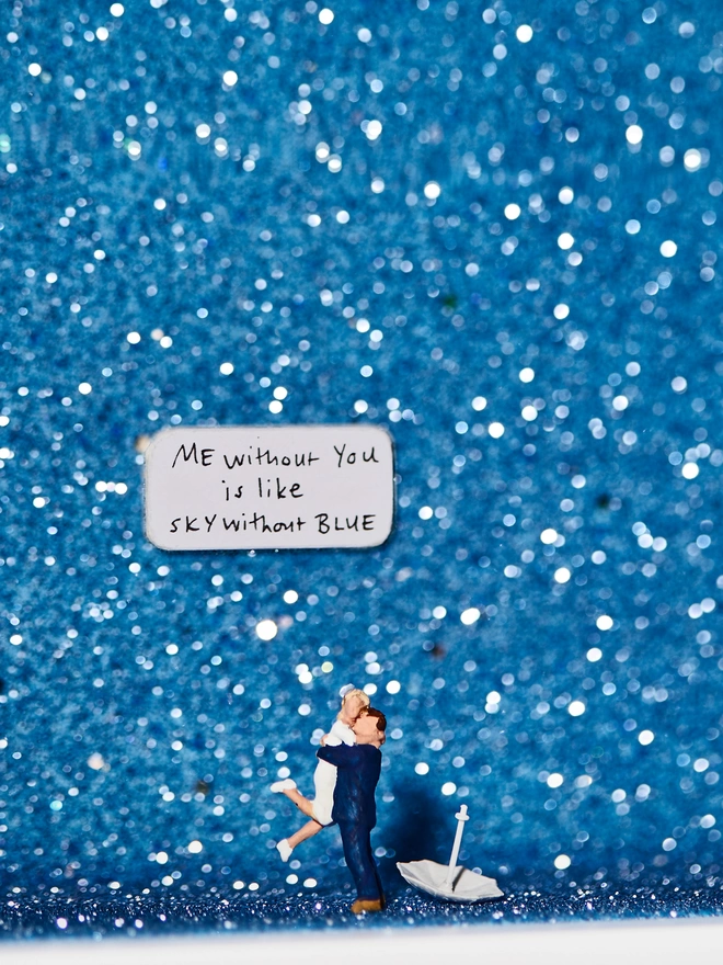Miniature scene in an artbox showing a tiny man and woman embracing against a dramatic blue glittery backdrop evoking a rainstorm. The words “Me without you is like sky without blue” are written on the back wall.