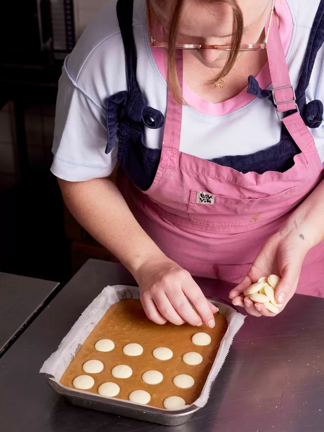 Founder Hetty wearing pink apron, adding white chocolate buttons to the blondie mixture in a baking tray within the bakery