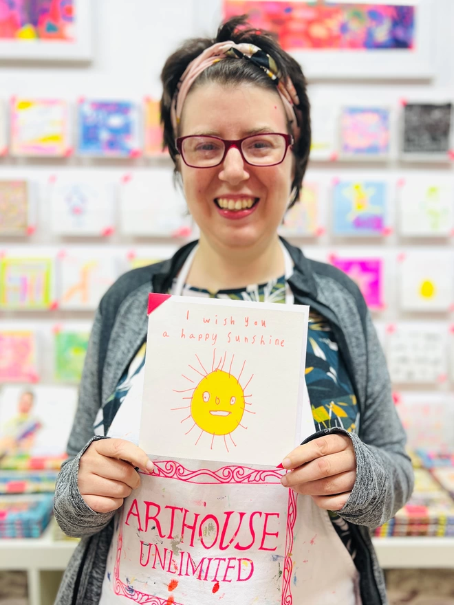 Artist holding I Wish You A Happy Sunshine card with a sketch of a smiling yellow sun outlined in orange
