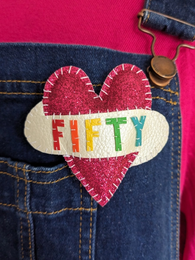 Pink glittery heart shaped brooch with FORTY spelt out in rainbow coloured lettering on a white scroll, pinned to blue denim dungarees