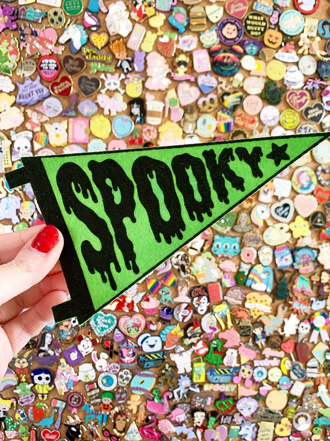 Image shows a hand holding a small felt pennant with the word ‘Spooky’ printed in black ink on bright green felt. The letters of the word are designed to look like the word is dripping. 