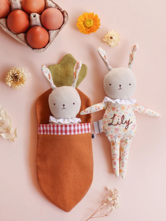 natural linen rabbit dolls with printed floral outfit and ruffled white lace collar with embroidered gentle face feature. One is snuggled in a linen carrot shaped sleeping bag.