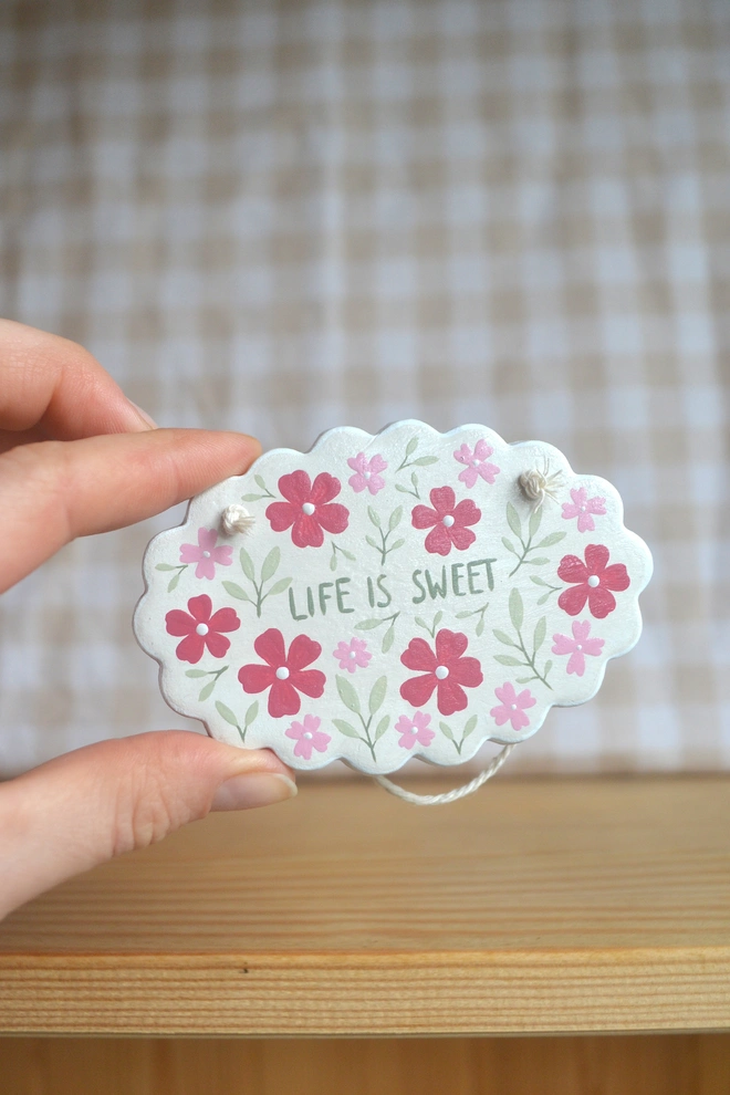 hanging clay sign with red, pink and green floral design reading 'life is sweet' in front of gingham background