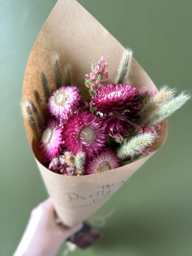 a bunch of dried flowers in bright colours wrapped in kraft paper held by a person in a mustard coloured jumper with their left arm against a green wall