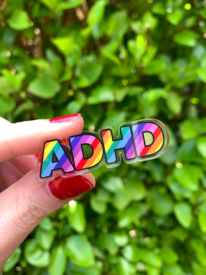 image shows an acrylic badge in the shape of the letters ADHD. the letters are filled with diagonal rainbow stripes.