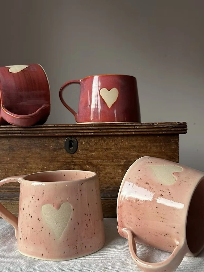 Heart Mug Strawberry Pink, pair of pink mugs on a white surface in front of another pair of red heart mugs, on a small wooden cabinet. 