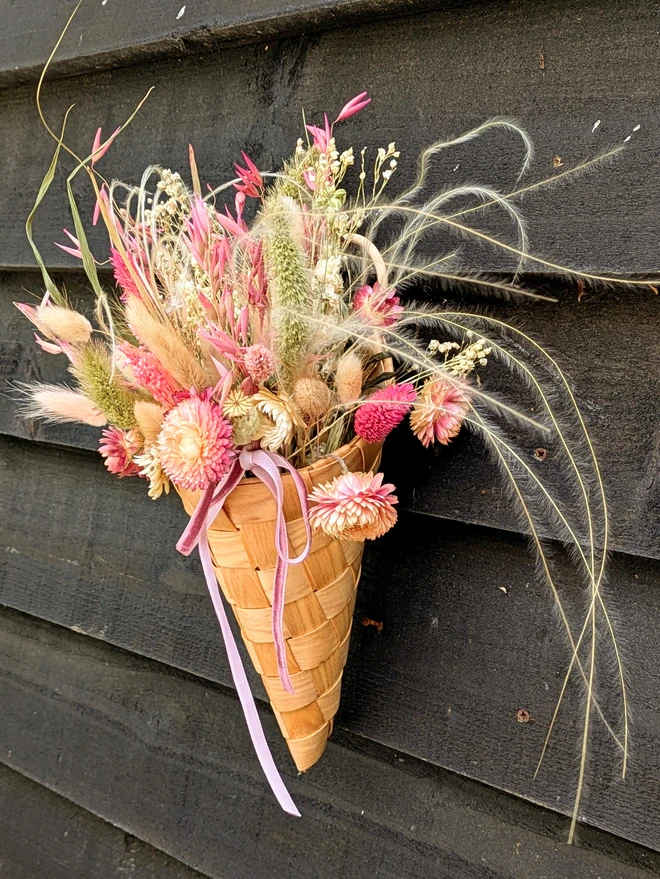 wooden woven cone basket with dried pink flowers hanging from a hook on a wall