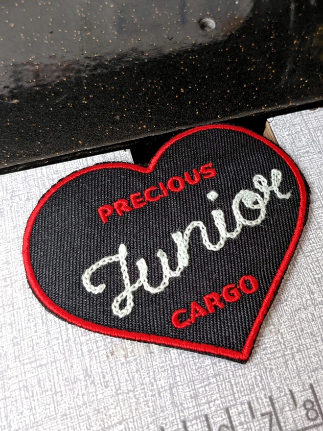 'Precious Cargo' Woven Embroidered Patch in Black & Red. The words 'Precious' in Red & 'Cargo' are digitally embroidered onto the patch, whilst the Chainstitch customisation reads 'Junior' in White thread, taking up the main space on the patch. The patch is resting on a sewing machine table. 