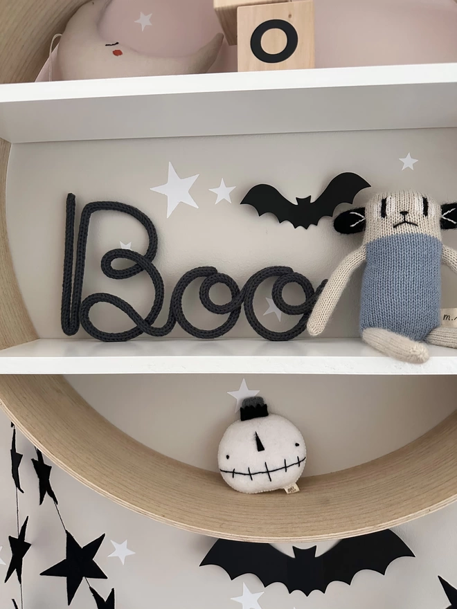 Knitted Word Sign - 'Boo' with a capital B sitting on a shelf next to a quirky soft toy character.
