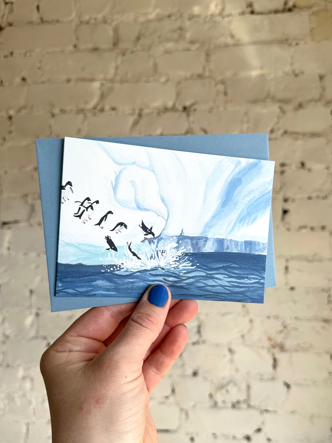  a greetings card featuring an illustration of an icy landscape with penguins diving into the sea with a blue envelope