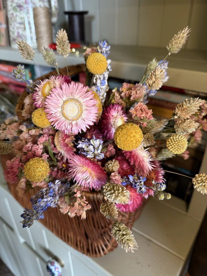 a bouquet of dried flowers in bright colours displayed in a wicker basket against a green painted dresser