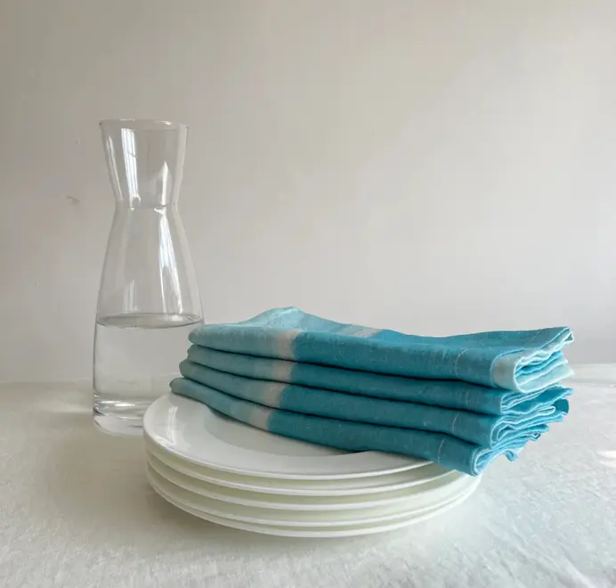 Azzurro Napkins (Set of 4). Light blue cloth napkin, styled on a white plate folded on a pile of plates. 