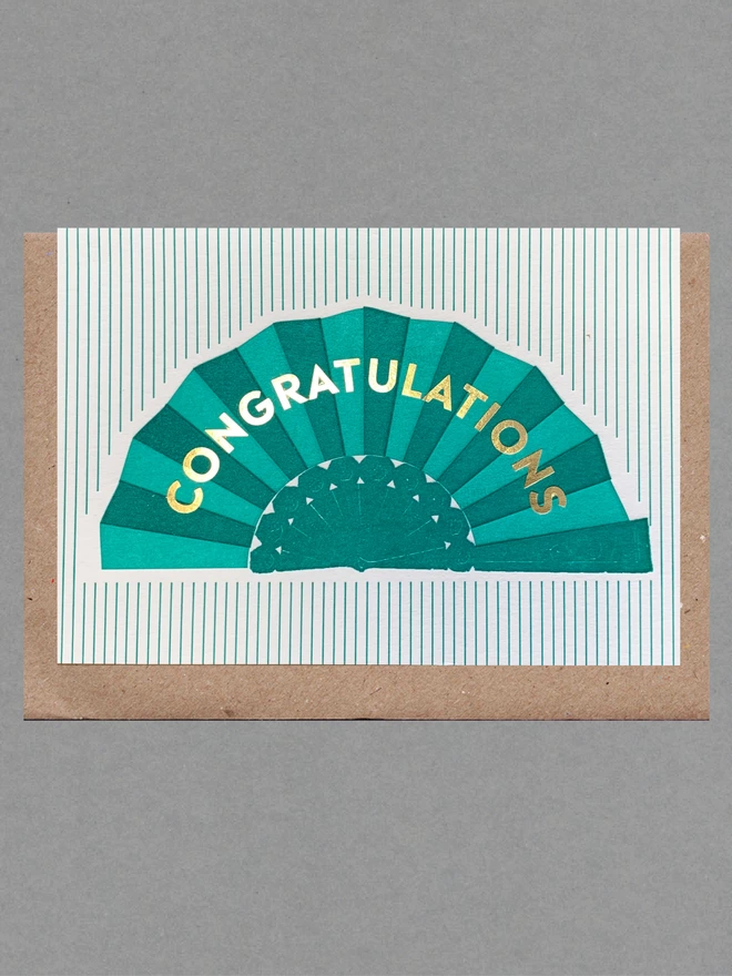 Green and white striped card with a green fan on it and gold text reading 'Congratulations' with a brown envelope behind
