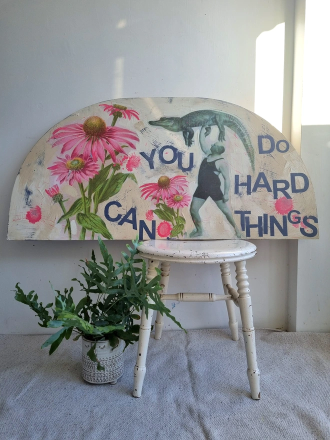 Art panel in a crescent shape made from a piece of vintage table top. Decorated with decoupage images of a man holding a crocodile aloft and pink flowers. The text reads 'you can do hard things'. The colours are cream with splashes of pink