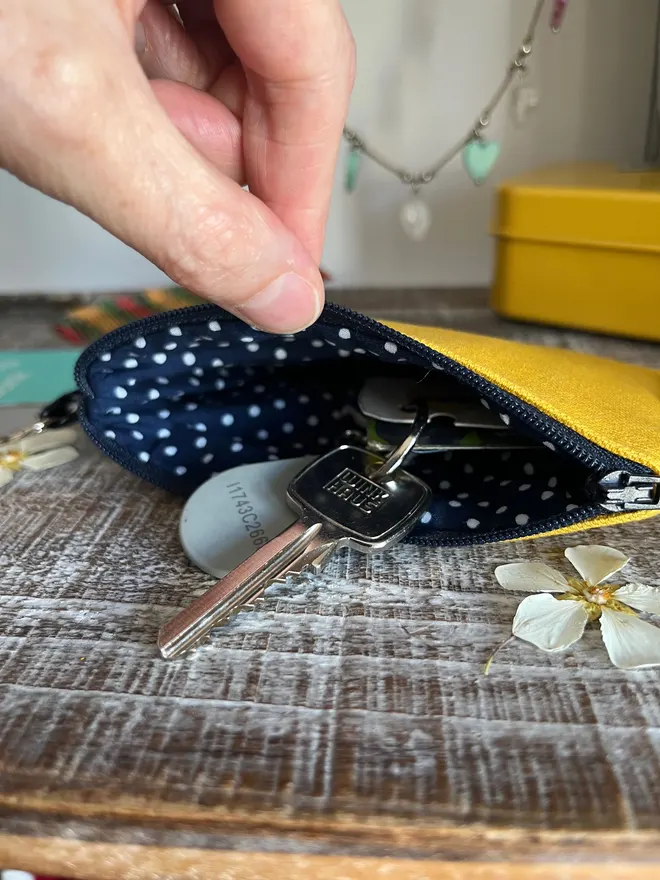Small yellow coin purse with zip open showing navy polka dot lining. The pouch holds a set of keys.