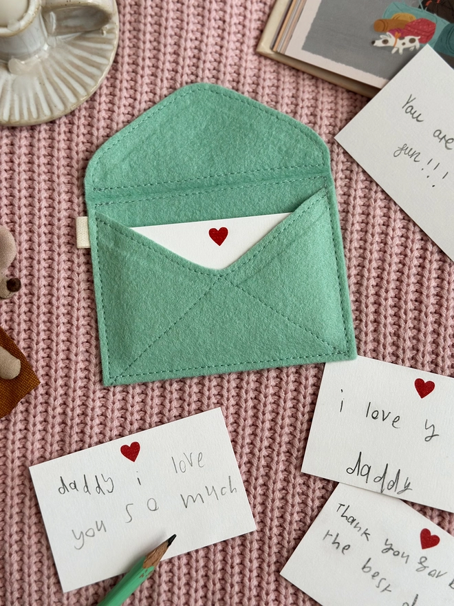 A handmade envelope lays open on pink fabric. Tucked inside are mini heart notecards, with several written cards beside it. Each written card has a note to Daddy written by children.