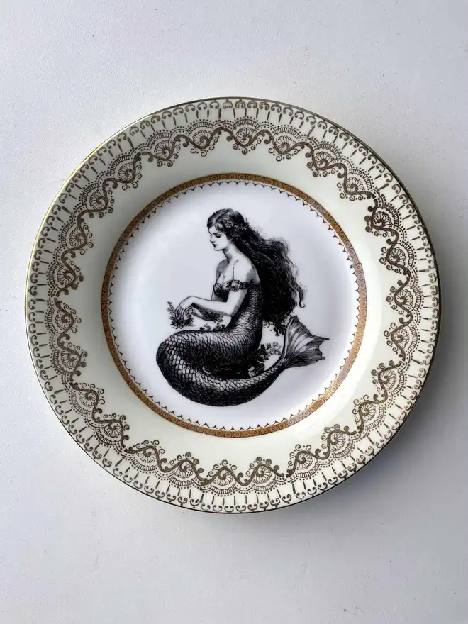 a vintage plate with an ornate border has a black and white vintage illustration of a mermaid in the centre
