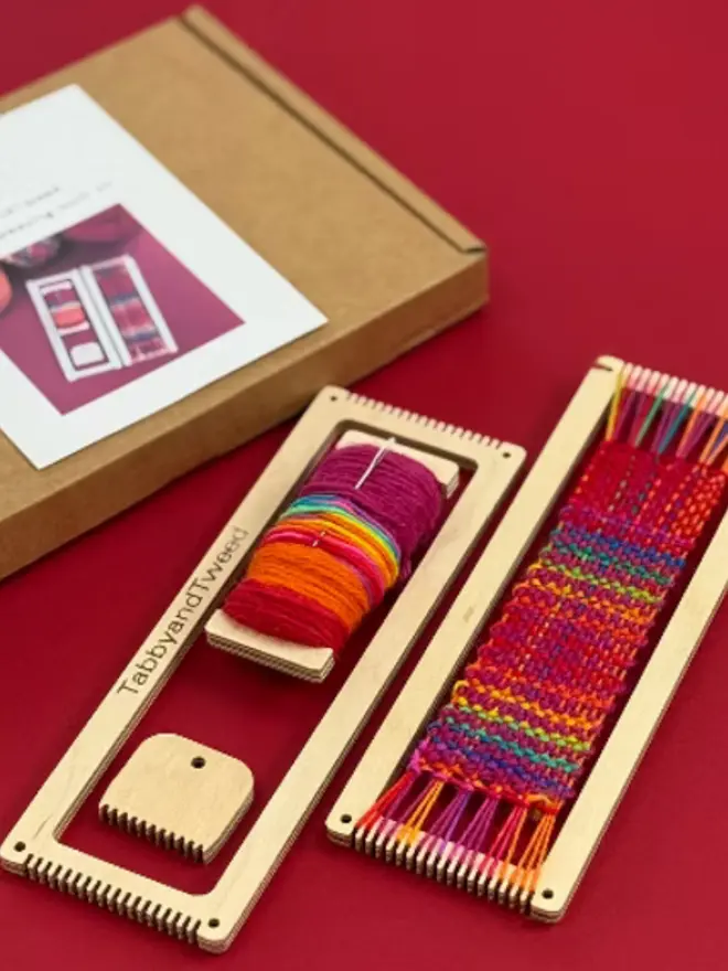 Rectangular beige looms, one woven with bright rainbow coloured yarns with a spool of bright yarn and wooden comb.