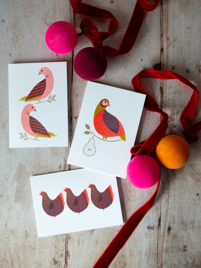 Bright and colourful, 3 maximalist feeling mini Christmas cards featuring partridge in a pear tree, two turtle doves and 3 French hens are scattered on a wooden table with jewel toned Christmas baubles and ribbons surrounding them