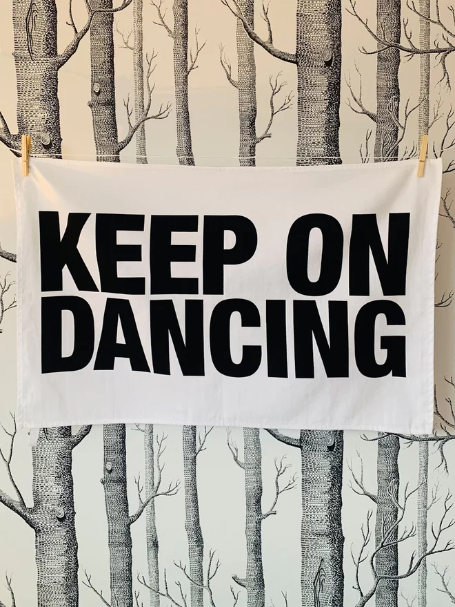 London Drying Keep on Dancing black screen printed text on white tea towel hanging washing line style with clothes pegs in front of white wallpaper that has black screen print pattern of silver birch trees