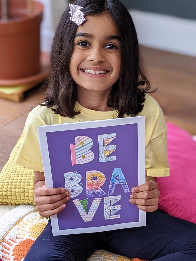 Young girl sitting holding an art print saying 'Be brave'