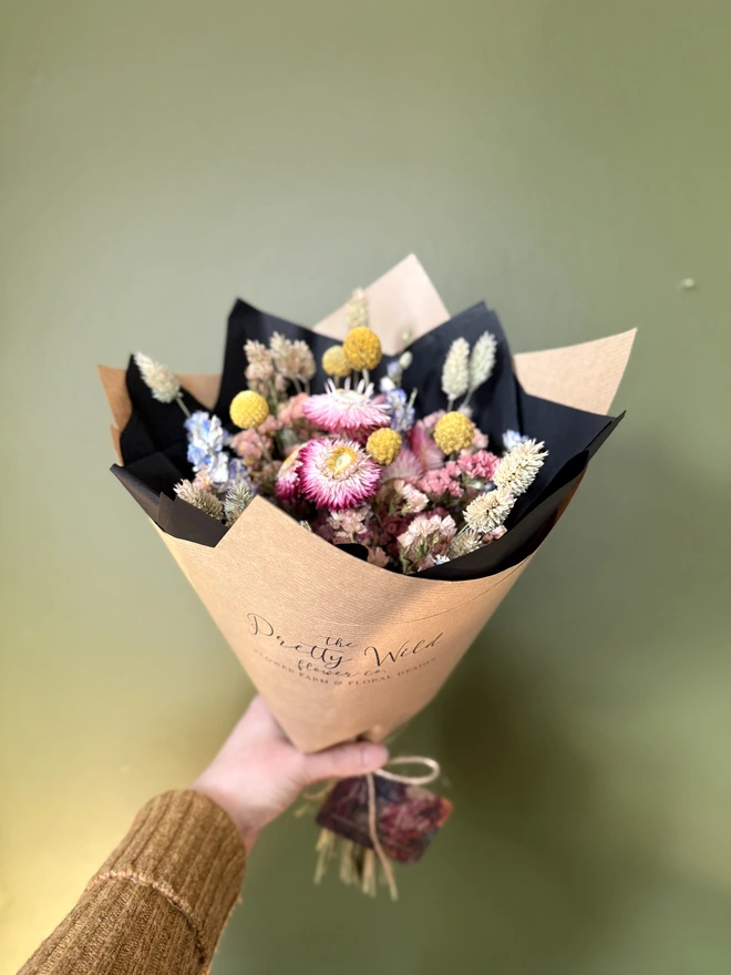 a bouquet of dried flowers in bright colours wrapped in black tissue and kraft paper held by a person in a mustard coloured jumper with their left arm against a green wall