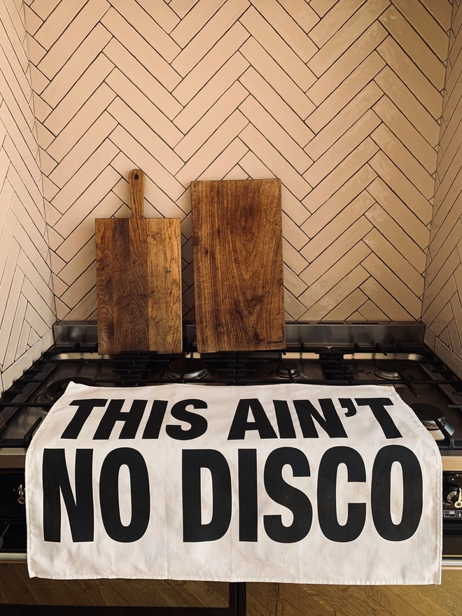 London Drying This Ain't No Disco black screen printed text on white tea towel laying on oven with 2 chopping boards in background
