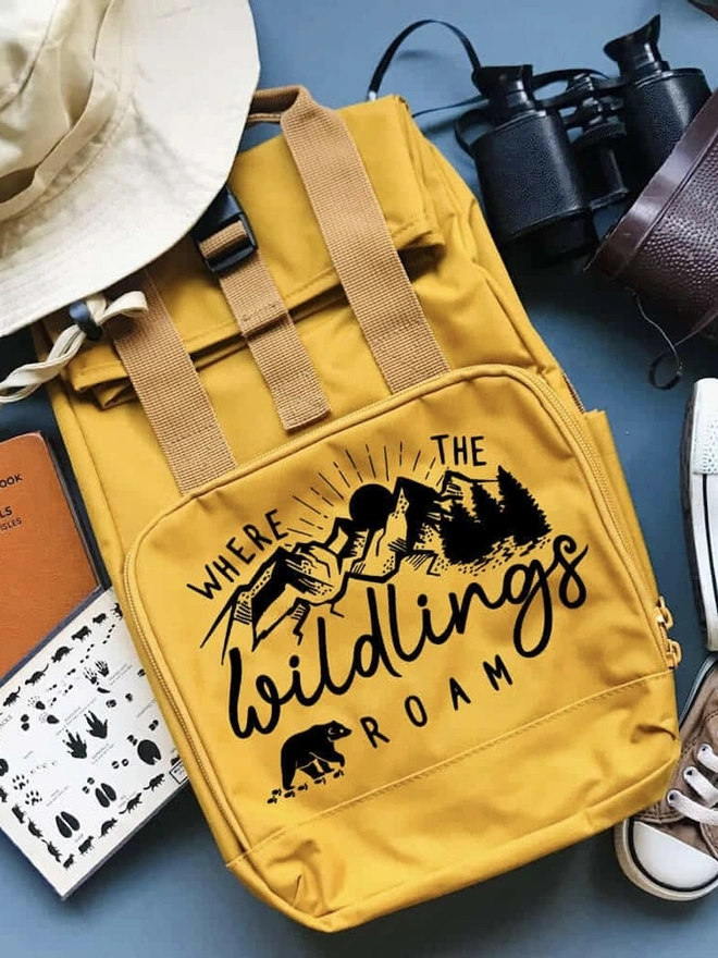 'Wildings' Children’s Recycled Roll Top Back Pack, a colourful backpack sitting on a blue backdrop accompanied by other items. 