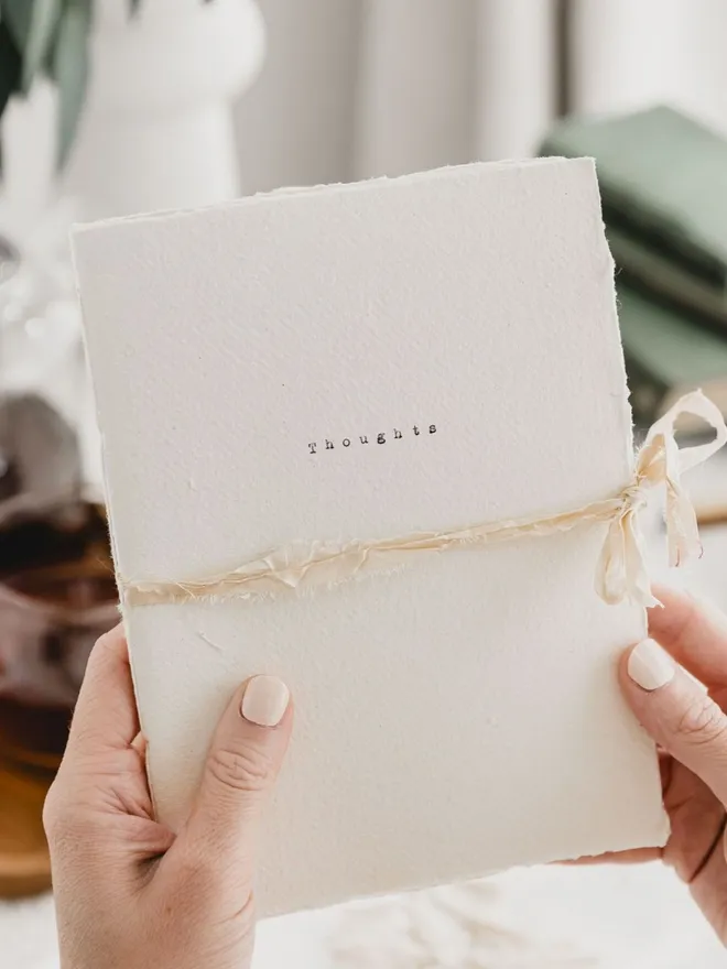 an off white journal with a deckled edge and rustic ribbon closure the word 'thoughts' has been typed on the cover