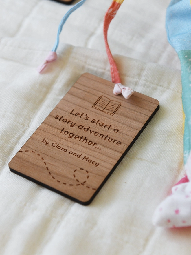 A wooden card lays on a white quilt. It has engraved words which read 'Let's start a story adventure together'.