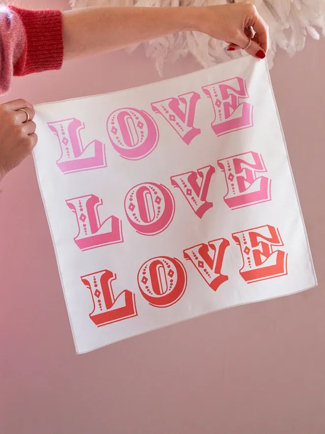 LOVE LOVE LOVE printed cotton handkerchief being held up against a pale pink wall with a white feather lightshade