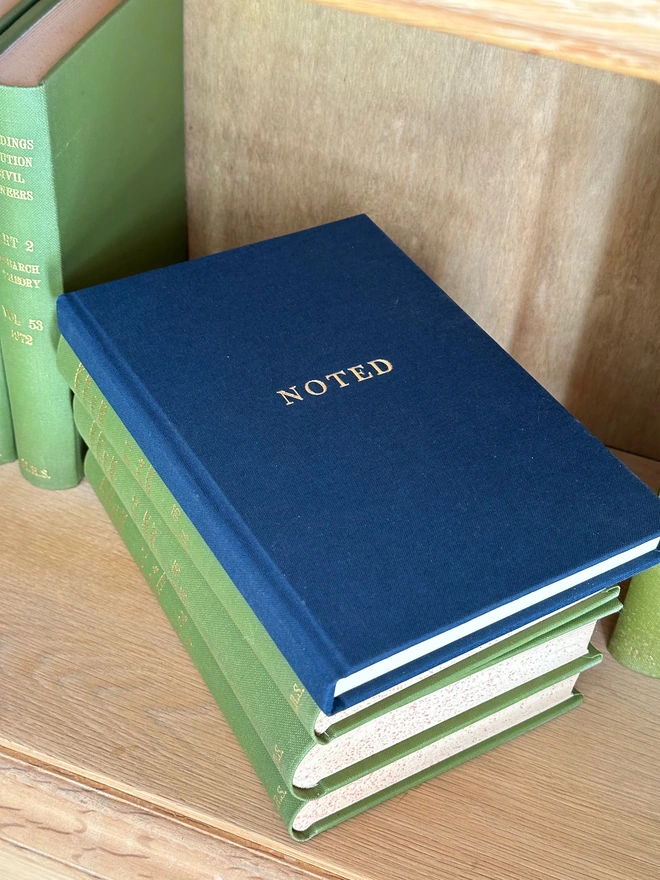 A navy linen notebook printed with 'Noted' in gold lettering sits on a pile of green books. on a bookshelf.