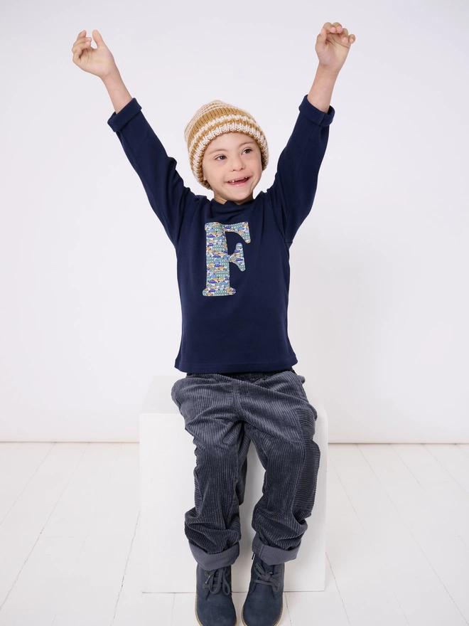 A navy cotton long sleeve t-shirt appliquéd with an initial in a vintage cars and busses Liberty print, worn by a 4 year old boy