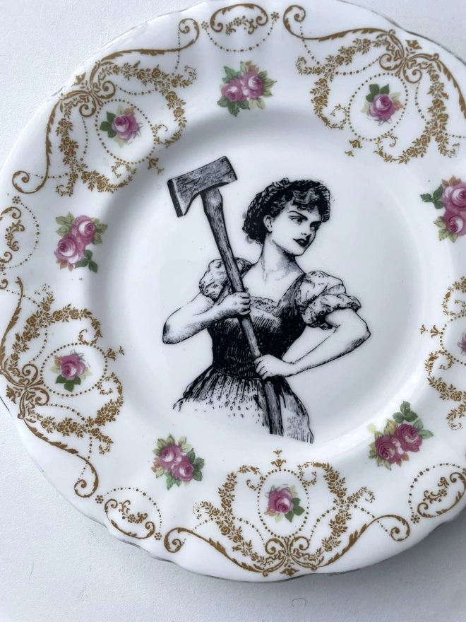 a vintage plate with a really ornate pretty border has a vintage black and white illustration of a beautiful girl in peasant clothes wielding an axe