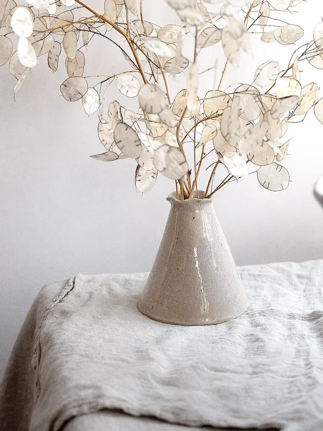 Maia Bud Vase In Sea Salt Glaze, with plant in it, placed on a table cloth against a white background. 