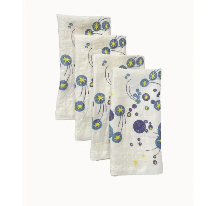 Solaris Napkins In White Or Blue. Set of four off-white cloth napkins with blue and yellow starburst and orbital patterns, stacked in a staggered arrangement.