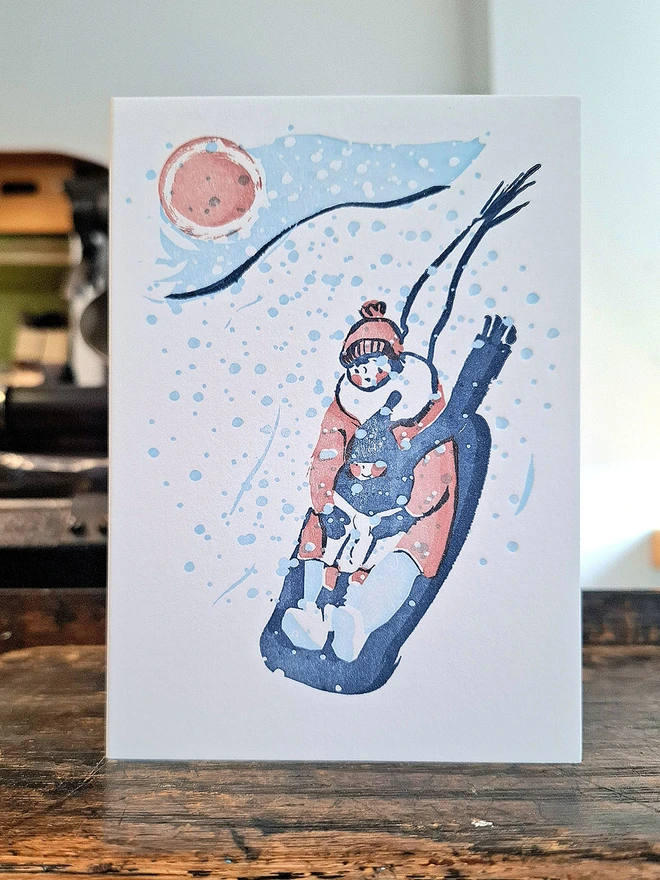 A handprinted greetings card standing open on a wooden surface.  The illustration on the card is printed in pink, light blue and dark blue, and is of a mother holding her young child as the sledge down a hill in snow.