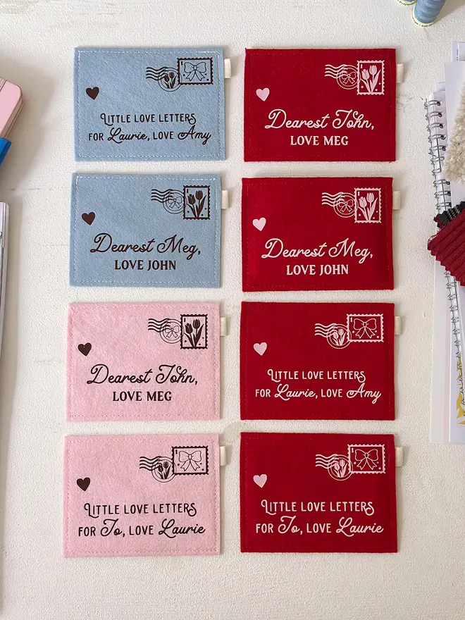 Eight handmade felt envelopes, each with a love letter theme, are arranged on a white wooden desk. Each envelope is personalised with two names.