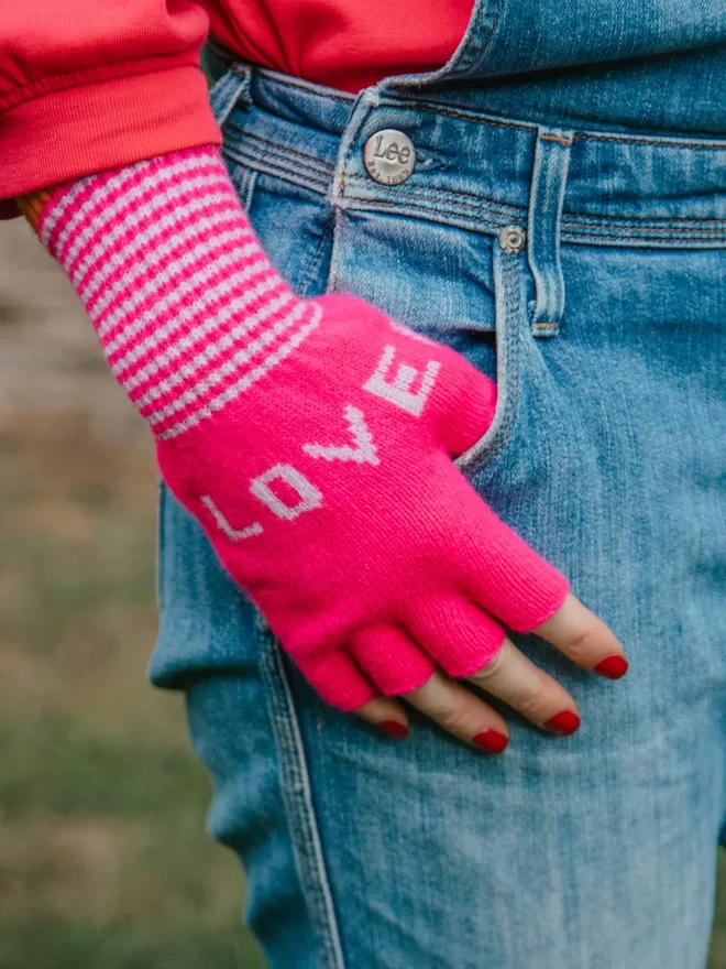 A bright pink fingerless knitted glove with the words LOVE and HOPE in lilac and a stripe cuff with orange edge detail. Made from 100% Lambswool with Wool spun in Yorkshire. 