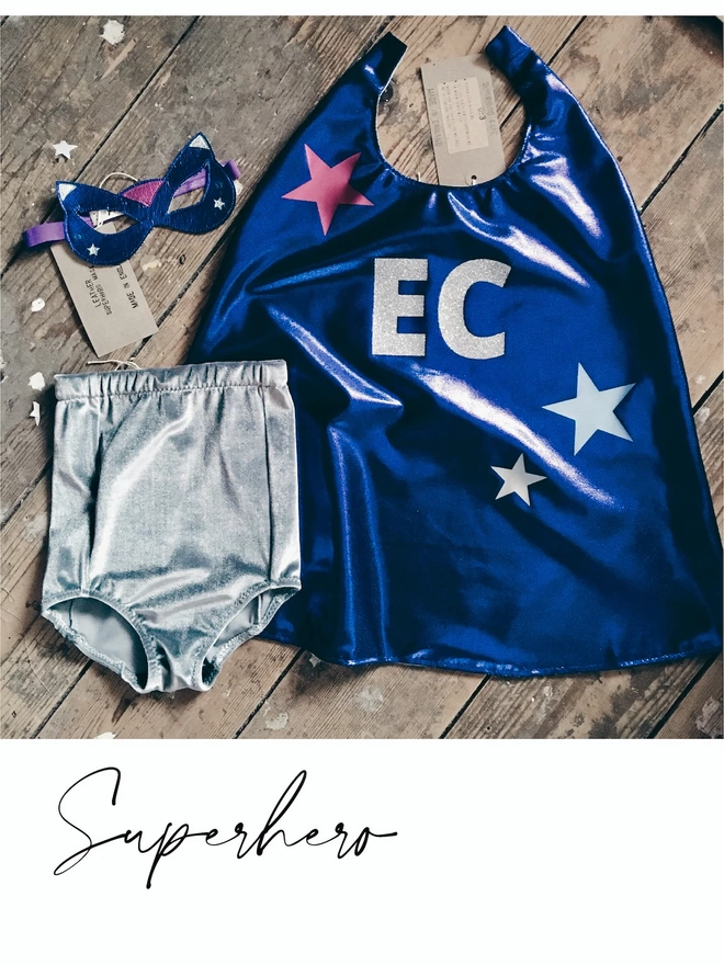 Flat lay of Blue Superhero costume placed on wooden floorboards. Sapphire blue superhero cape with silver glitter initials and star deign, Blue leather superhero 'cat' mask,  grey velvet shorts