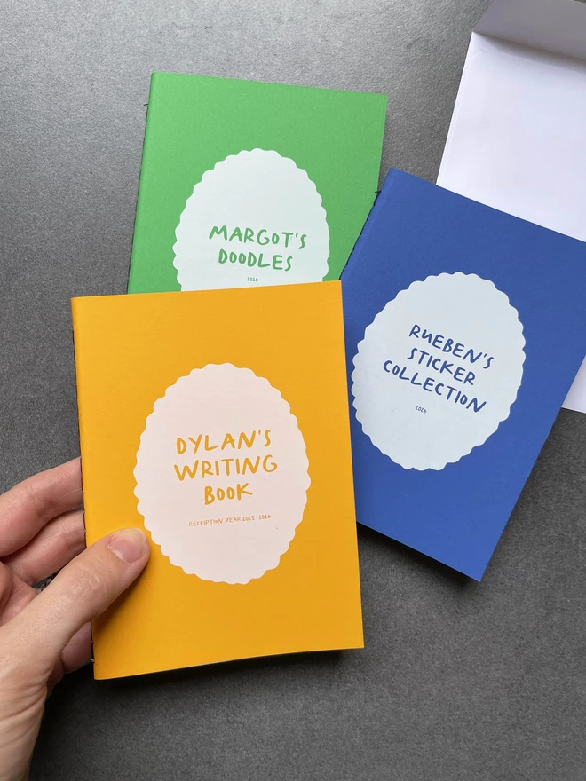 Three personalised brightly coloured pocket size notebooks on a dark worktop, one of the books says Reuben's sticker collection, one says Margot's Scribbles and the other says Dylan's writing book.