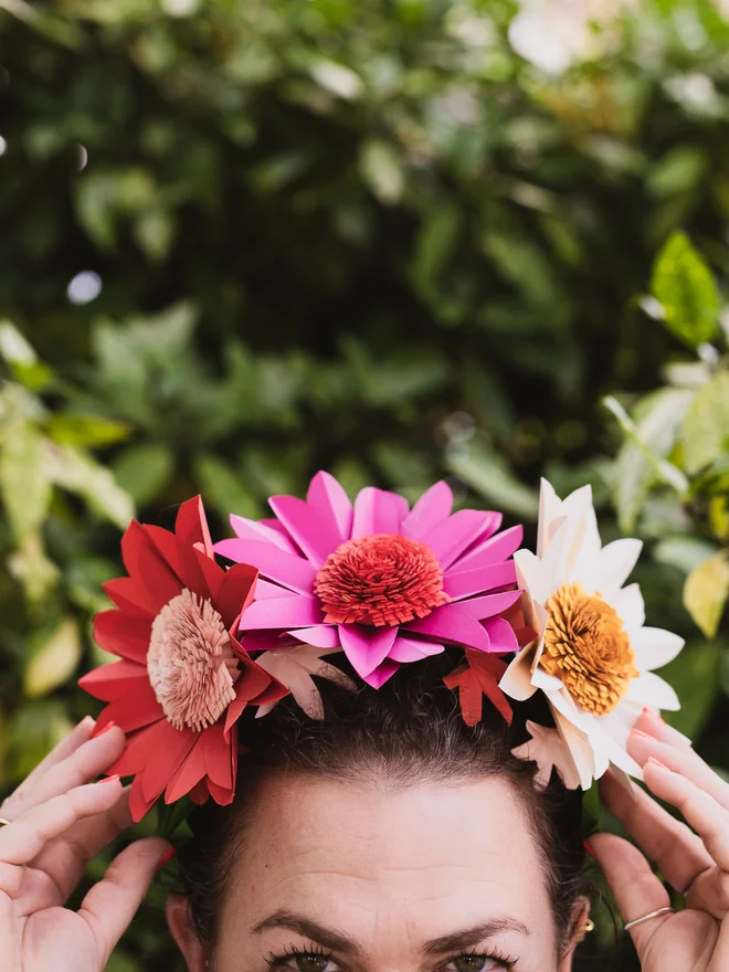 Woman with paper flower crown on head, with bushes in the background.