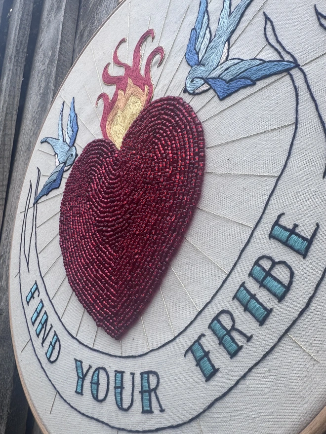 Embroidery hoop containing embroidered swallows a red beaded heart gold rays and a banner saying find your tribe 