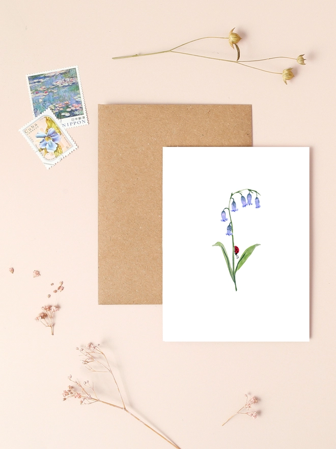 An image of a mini, rectangular white greetings card with an illustration of a bluebell flower with a tiny black and red ladybird crawling up the stem. The mini greetings card is placed on top of a rectangular brown kraft envelope.