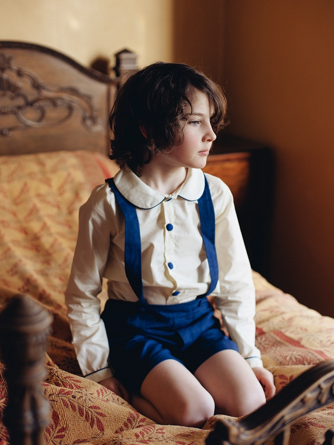 A boy sits on a bed wearing a cream shirt with navy piping and buttons and navy shorts with braces