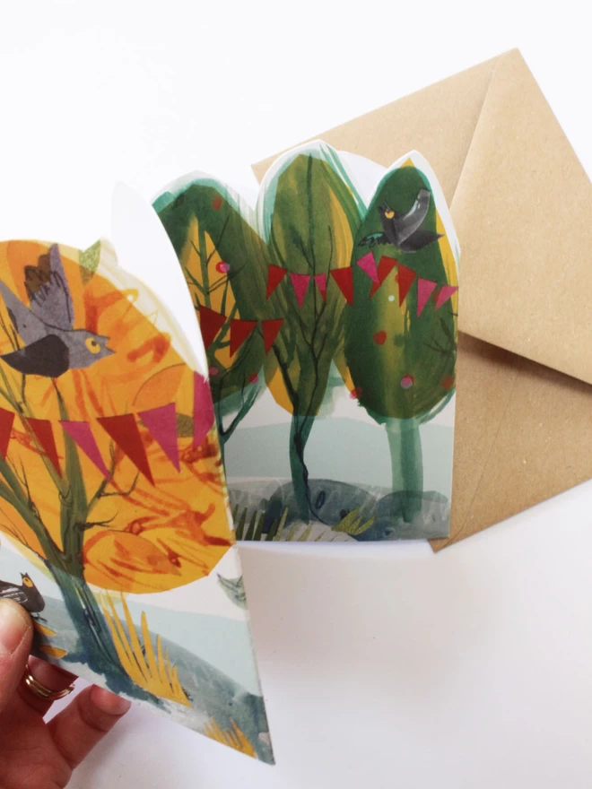 Hand pulling out colourful  concertina card of illustrated yellow and green trees